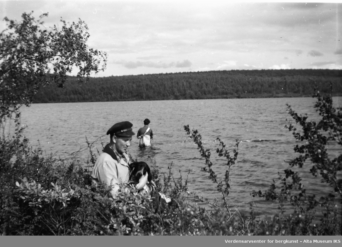 En mann sitter på elvebredden med en hund i fanget, En annen man står ut i elva å fisker.