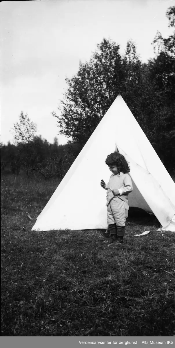 Et lite barn med fyldig og krøllete hår står foran åpningen på et telt, med skogen i bakgrunnen.