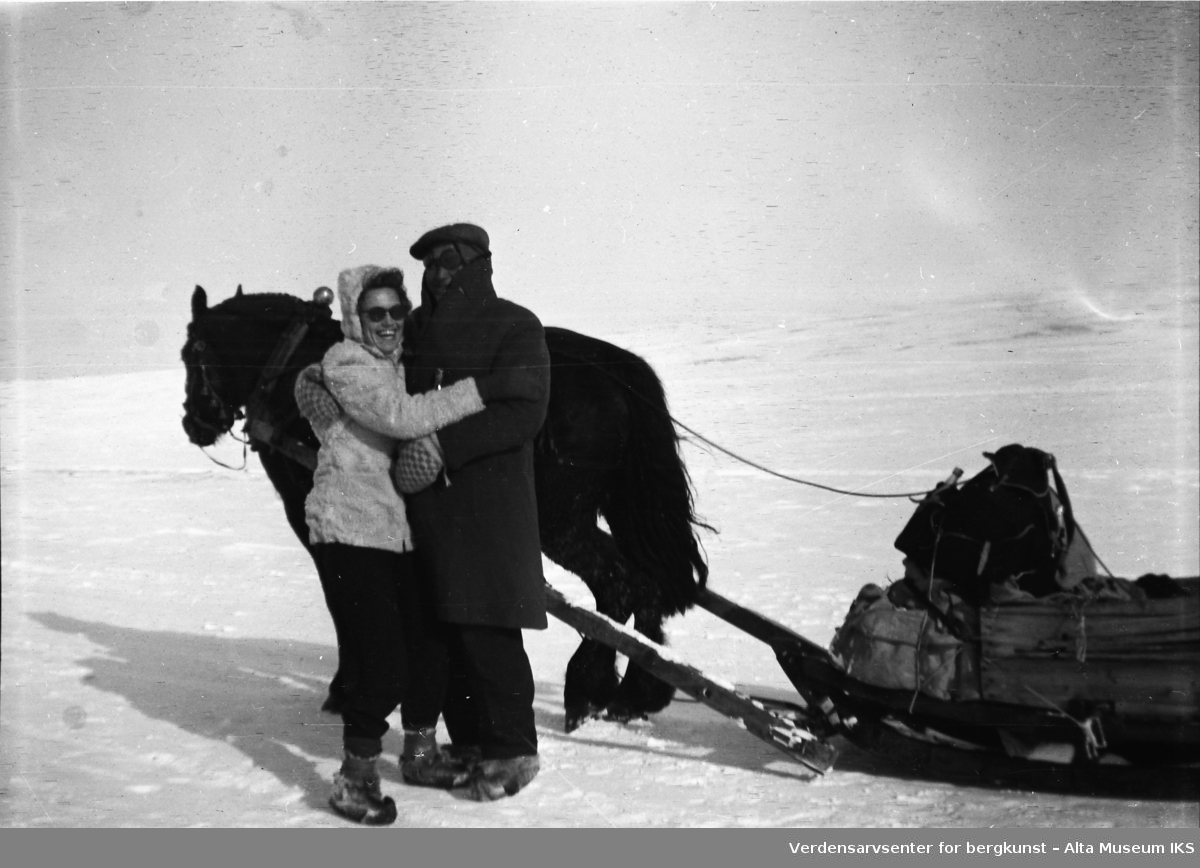 En mann og en kvinne står foran en hest med slede på fjellet. De holder om hverandre. Snølandskapet er tom og flatt bak dem.