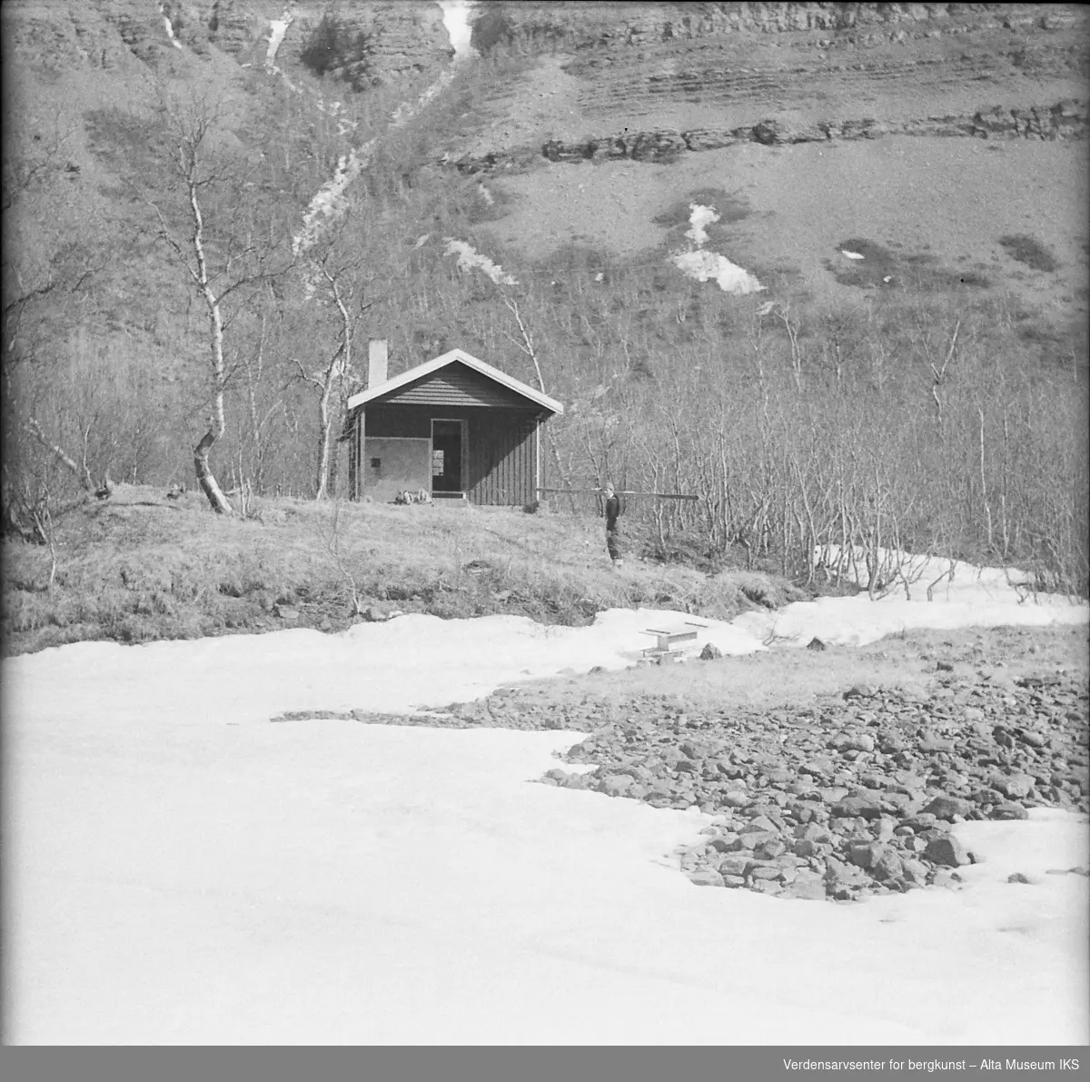 En mann bærer en planke til en hytte i skogen. Snøen har ikke smeltet helt, og bak skogen ser man foten av et fjell.