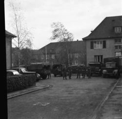 Fem uniformerte menn står foran militære lastebiler i en gat