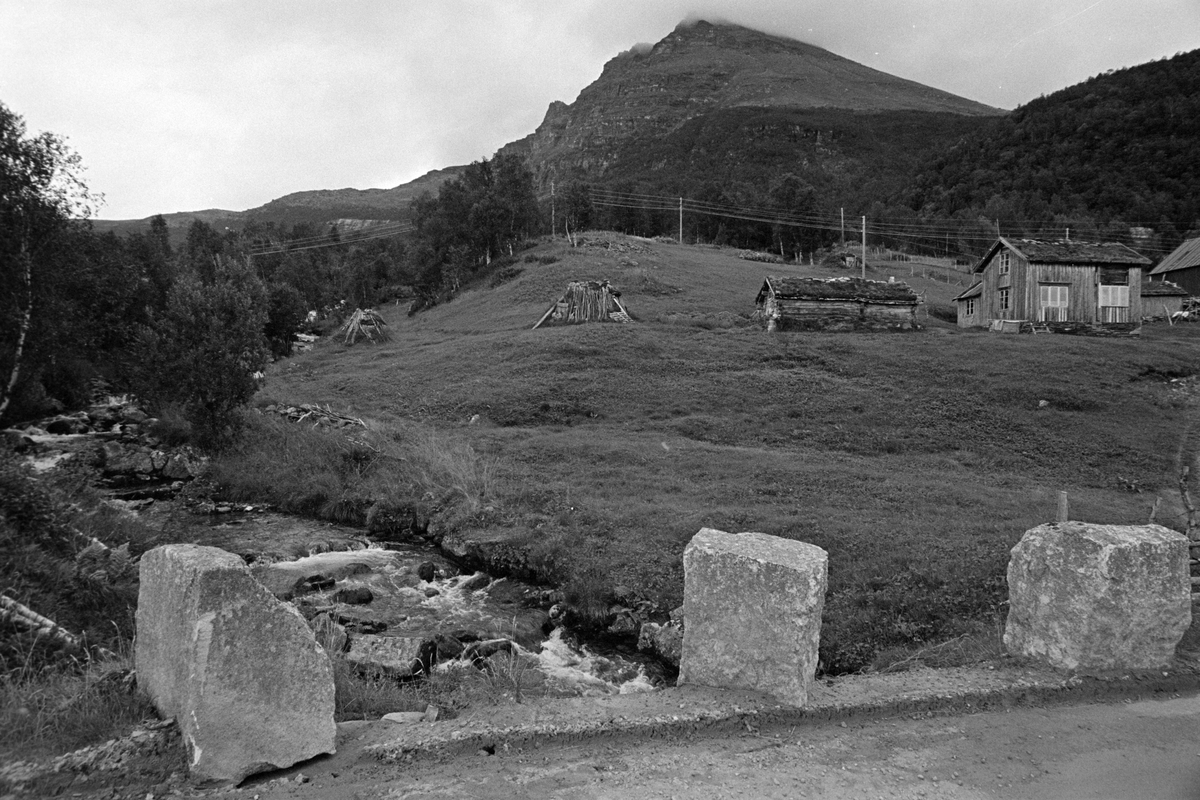 Gamme og gårdsbebyggelse ved elv, fotografert med kantstein ved veien i forgrunnen og fjell i bakgrunnen.