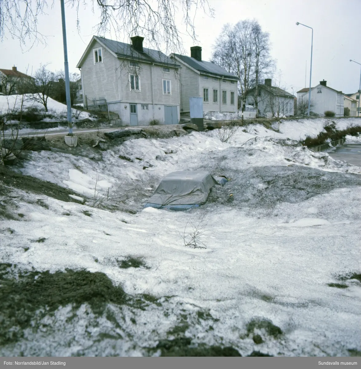 En översnöad bil tinar fram på en parkering på Södermalm våren 1977. Träkåkarna som syns på Nybrogatan och Nygatan finns fortfarande (2015) kvar.