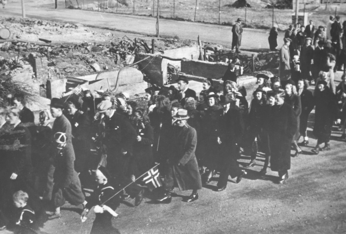 17.mai-tog i Kirkenes etter krigen. Voksne i hatter og frakker. I bakgrunnen ser vi ruiner etter bombinga.