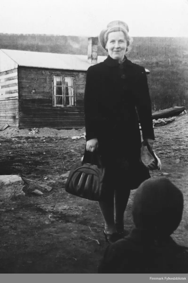 Lærer Gjerdrud Betten står foran en provosorisk bolig i Nesseby i gjenreisningstiden. Hun er kledd i en mørk kappe / jakke, basker og har en dameveske i handen. På forgrunnen foran henne står en liten gutt med ryggen til kamera.