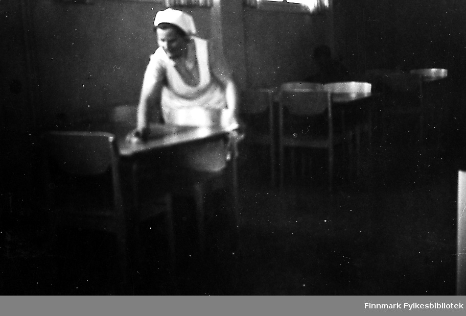 En kvinner tørker bordene på skolekjøkkenet i Seida skole. Hun har på seg en hvit forklær og tørkle.