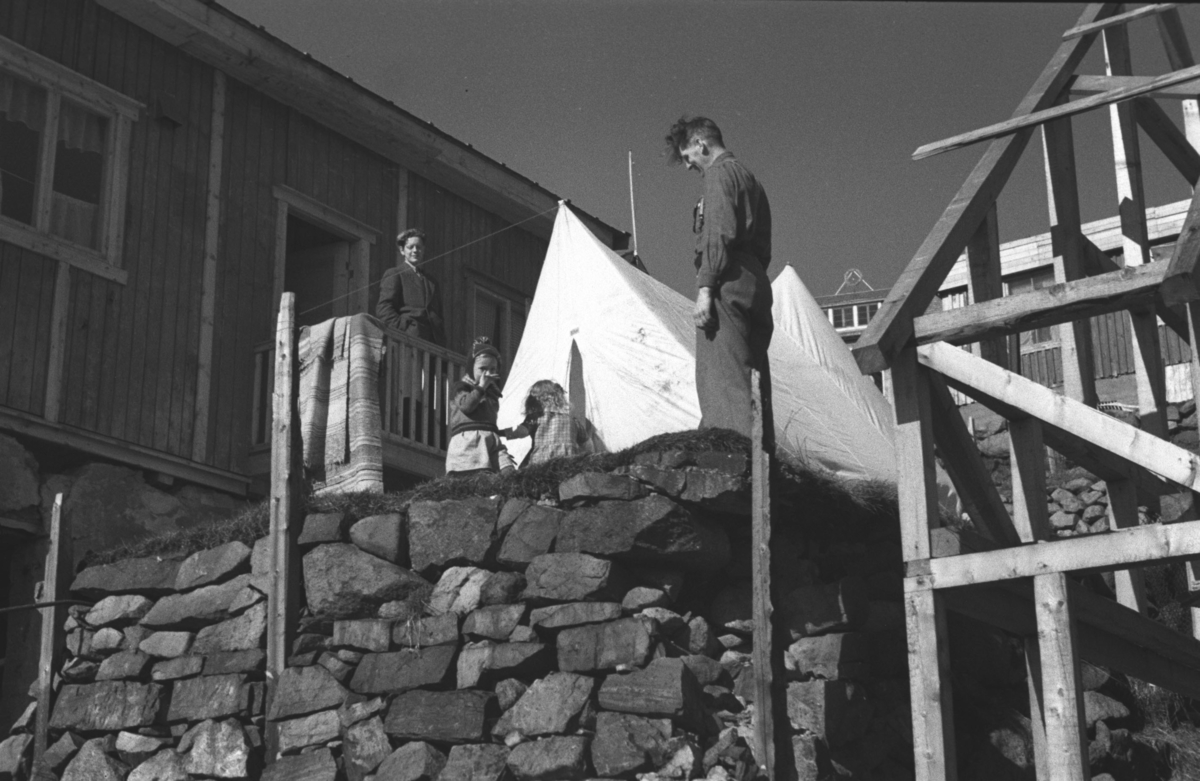 To menn med to små barn utenfor et telt som er slått opp utenfor et gjenreisningshus i Honningsvåg.

Arkitekt Ola Hanche-Olsen arbeidet ved Brente Steders Reguleringskontor i 1946. Hovedadministrasjon for gjenreisning av Nord-Troms og Finnmark ble lagt til Harstad og fikk navnet Finnmark kontoret. Landsdelen Nord-Troms og Finnmark blev oppdelt i syv distrikt med hver sin administrasjon. Honningsvåg, distrikt IV, skulle betjene Nordkapp, Lebesby, Porsanger og Karasjok kommune.

Ola Hanche-Olsen har tatt bildene. Han var født 13. mars 1920 i Borre, død 11. februar 1998 i Gjettum. Han hadde artium fra 1939, arkitekteksamen fra NTH 1946 og arbeidet deretter ved Finnmarkskontoret 1946–48 før han etablerte egen arkitektpraksis. Han debuterte som barnebokforfatter i 1974 med lettlest-boka "Knut og sjørøverne", og skrev i alt 12 bøker. 

Han var XU-agent 1944-45, og var også en aktiv fjellklatrer og friluftsmann. XU var den største og viktigste allierte etterretningsorganisasjonen i det okkuperte Norge under andre verdenskrig. Det meste av XUs virksomhet ble holdt hemmelig til 1988. Ola var gift med Solveig Hanche-Olsen (f. Falkenberg); de fikk 3 barn, blant dem matematikeren Harald Hanche-Olsen. 


