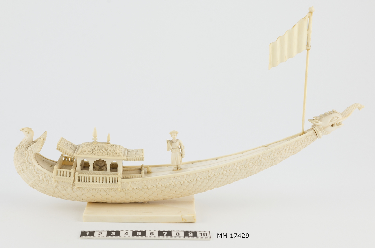 Modellbåt av elfenben föreställande kinesisk båt. Båtens skrov har formen av en bete med en smala, spetsiga änden till akter, som höjer sig högt över vattenlinjen. Fören är utformad som en påfågel med korta vingar samt kroppens och stjärtens fjädrar som ornament längs hela fartygets utsida. Den avsmalnande aktern är längst ut försedd med ett huvud föreställande ett fantasidjur med snabel, huggtänder - som blandning mellan elefant och drake. Närmast fören är en överbyggnad med tak och baldakiner, inuti sitter figur med korslagda ben. Mitt på däck står en man mot babord, hållande en stav lyft i höger hand. I aktern sitter en flagga på en stång. Längs kanterna på däcket är borrade hål, troligen avsedda för placering av figur som ror. Båten är placerad på rektangulär platta 60 x 90 mm.