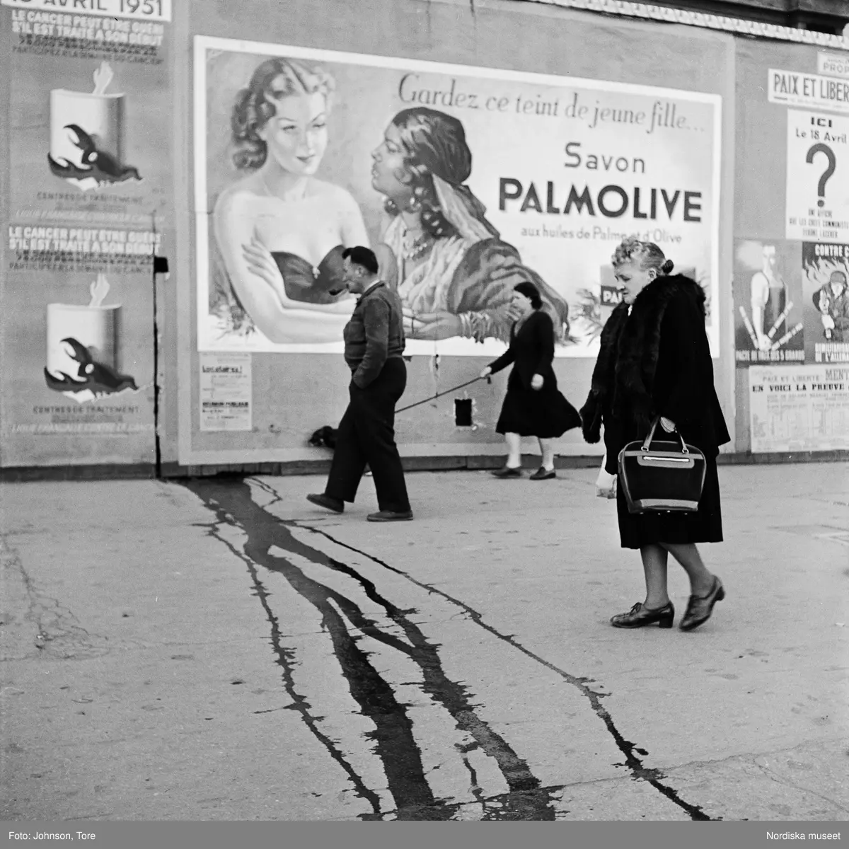 Paris. Fotgängare på gatan, reklamaffisch för Palmolive på fasaden i bakgrunden. Vatten (?) i rännilar på gatan.
