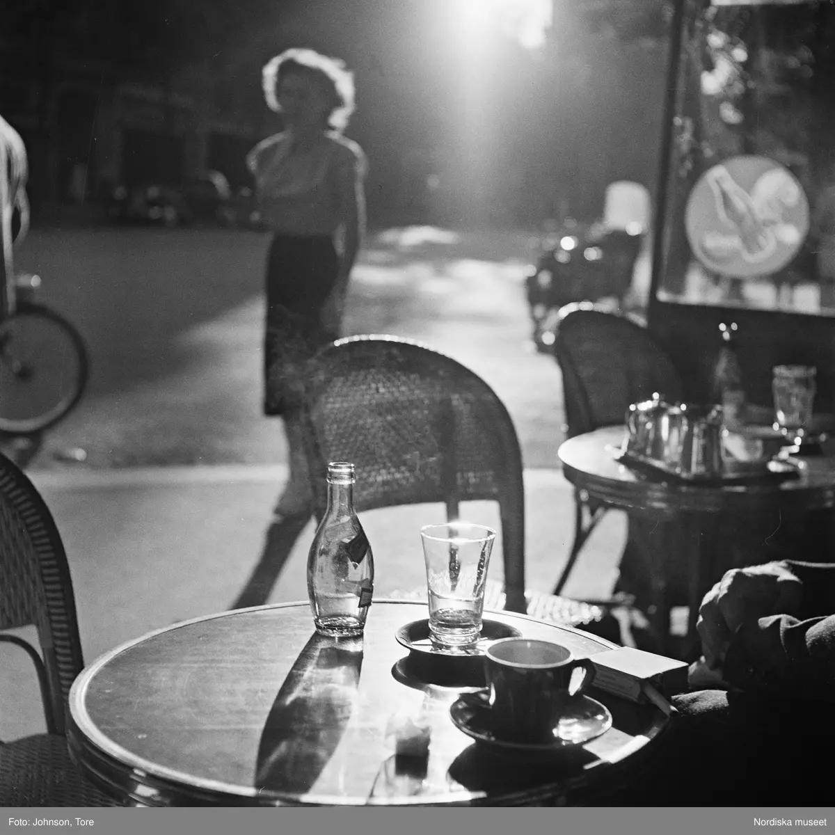 Paris. Ett cafébord på gata i motljus. Konturerna av en kvinna på gatan i bakgrunden.