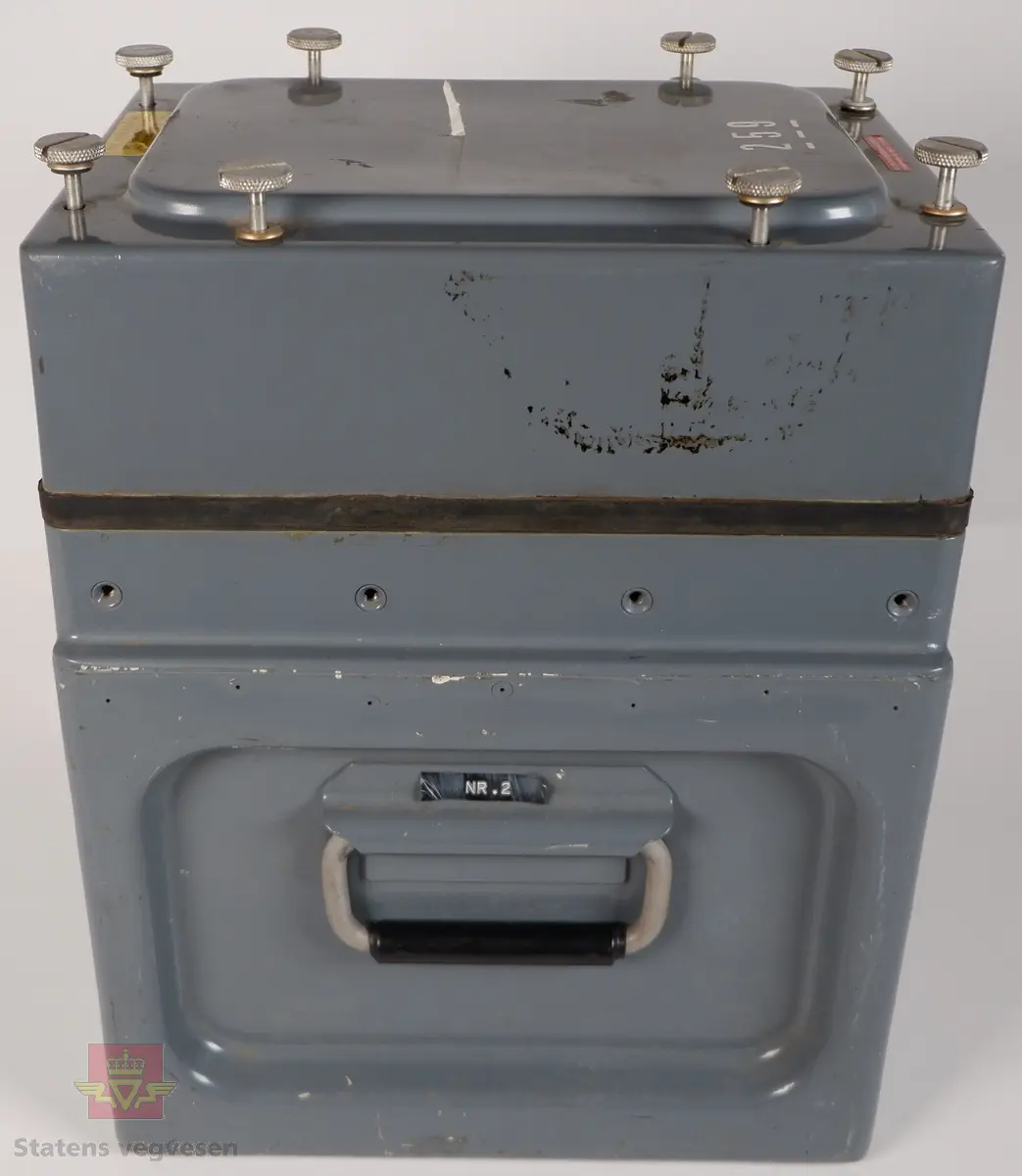 Vibrasjonsmåler i kasse av grålakkert metall. Kassen har lokk som er festet med skruer, og har påskriftene THIS COVER SCREW TO BE RELEASED BEFORE TRANSPORTING BY AIR, 259 og KONTOR FOR FJELLSPRENGNINGSTEKNIKK FORSKNINGSVN. 3B - OSLO 3 - TLF. 46 98 80, På undersiden av lokket er det teipet på et maskinskrevet ark som har påskriften VAKTHUND Ved kontroll: 1. Før strimmelen tas ut slåes inn dato og tid. 2. Vakthund slåes av og på (for å nullstille tidstellingen) 3. Slå inn dato - tid og instrumentnr. på strimmelen som blir sittende. 4. Husk å slå av bryteren på regnemaskinen. Rystelsesmåleren har to punkter for tilkobling med kabel, og kalkulator med skriver merket MONROE 10. Kalkulatoren har en håndskrevet klistrelapp som er påført datoer for bytte av batterier.
