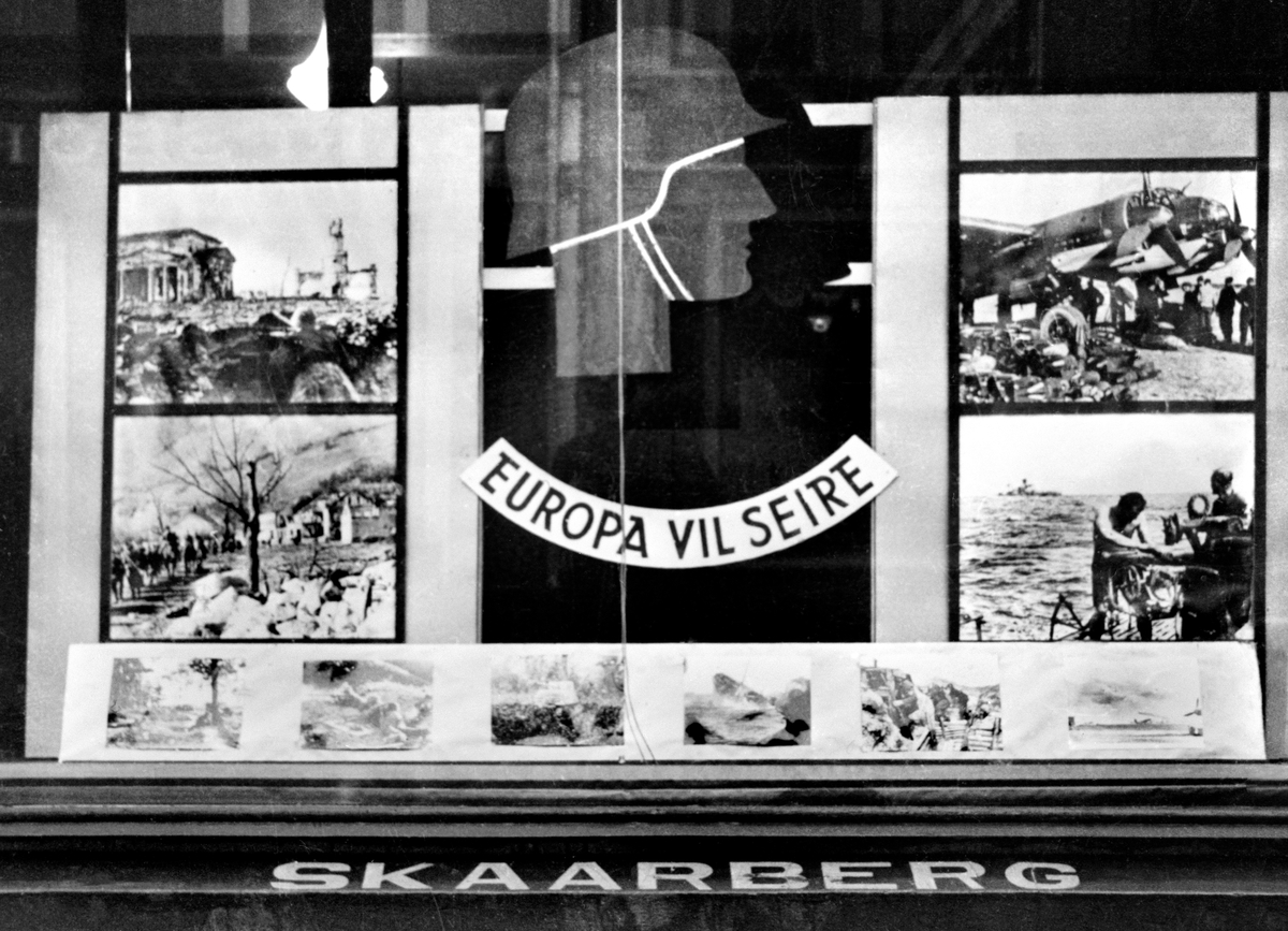 Plakater og utstilling med propaganda fra okkupasjonstiden, 1941-45, utstilt i butikkvindu i Sarpsborg, i St. Marie gate 105, i manufakturforretningen til Einar  Skaarberg A/S.
"Europa vil seire".

Butikkfasaden til Sarpsborg Maskinforretning speiler seg i vinduet, St. Marie gate 86.