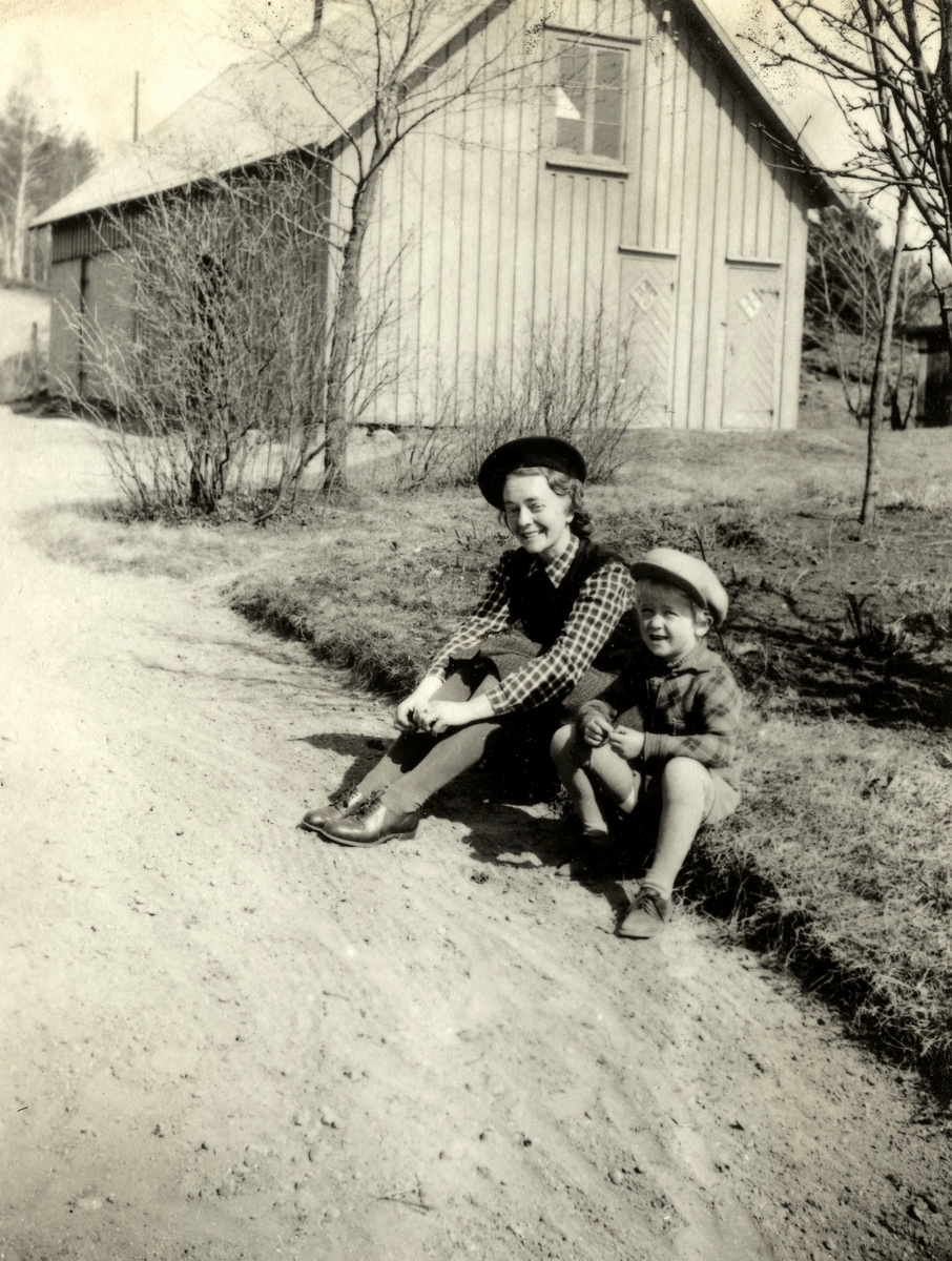 Rosa Krantz (1912 - 1994, gift Pettersson) och en liten pojke sitter vid vägkanten utefter Tulebovägen, 1930-tal. I bakgrunden ses vedboden som låg vid föräldrarnas personalbostad i Stretered.