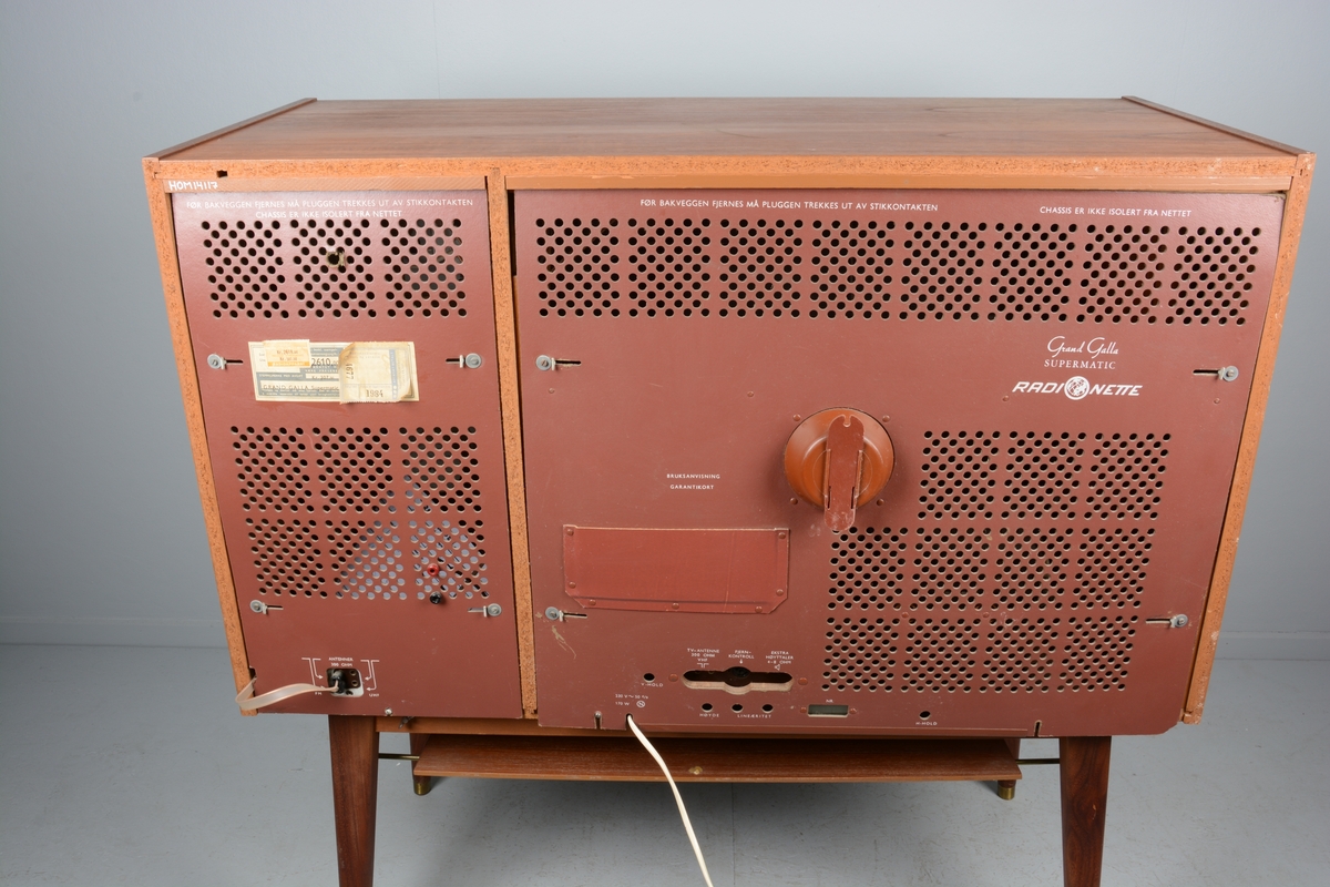 TV/fjernsyn samt radio monterte i kabinett med skyvedører for skjuling av fjernsynet når det ikkje var i bruk. Beteningsknappar og elektrisk kabel. Kabinettet har liggande rektangulær form.