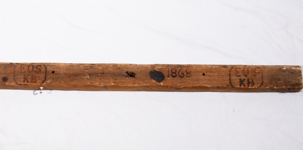 Stämpel på en träplanka. Har två stämplar på sig. Stämpeln är från Trummelsberg. Stämpeln är ett stort "B" som sitter ihop med ett stort "I". Har texten "EOS KB 1868" på sig.