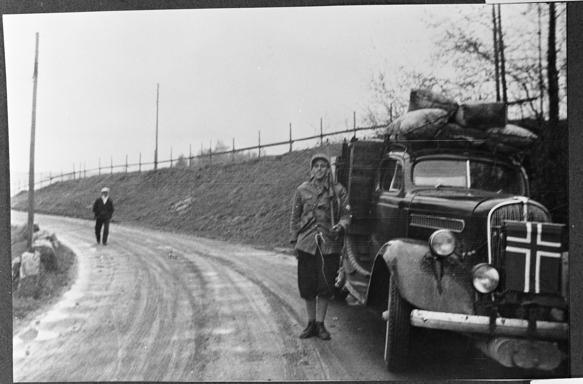 Kai Taralrud med flaggsmykket bil i nærheten av Totenvika kirke i maidagene 1945. Ole Engelund kommer spaserende i bakgrunnen.