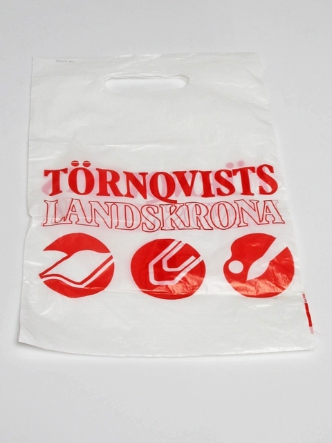 Plastpåse med reklamtryck: Törnqvists Landskrona. Påsen är i vit plast med rött tryck. Påsen pryds av runda bilder med stiliserade motiv. På ena sidan bild av en person som sitter vid ett skrivbord, på andra sidan tre bilder med respektive en uppslagen bok, ett gem och en palett med pensel.