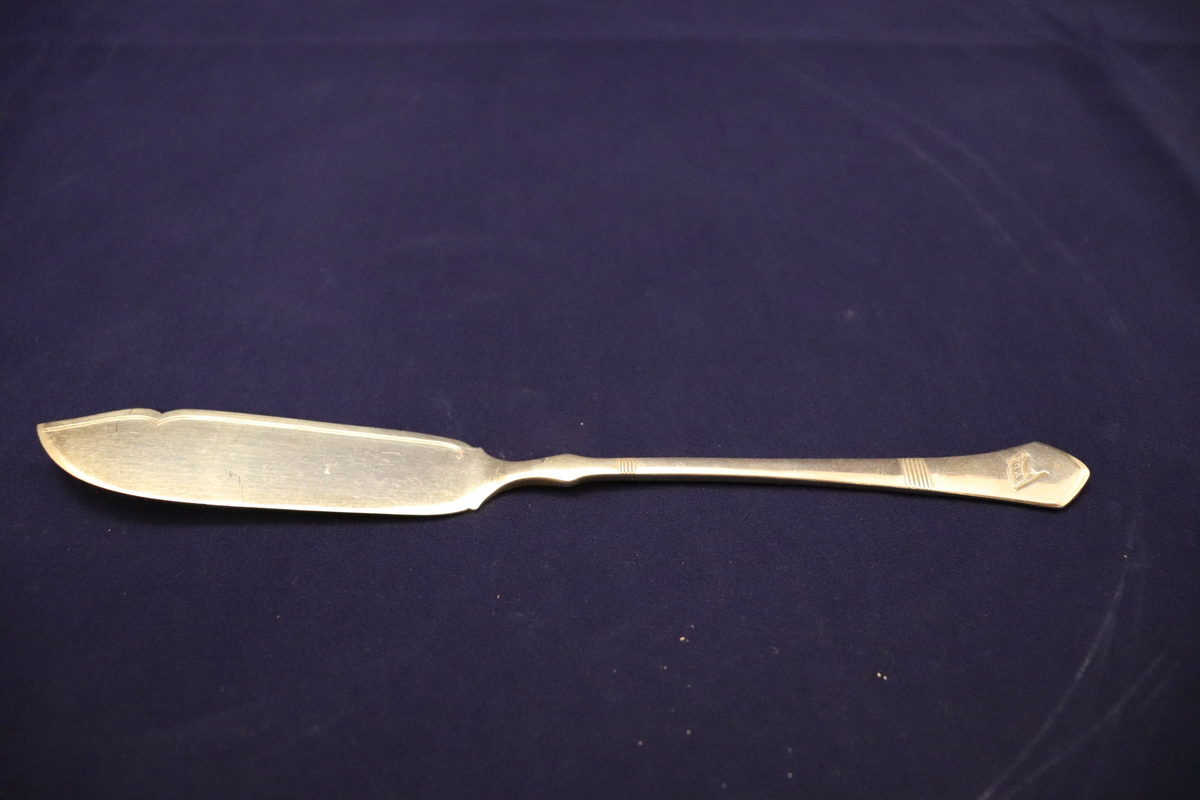 Kniv brukt til servering av fisk, trolig ombord i MS "Vesterålen"(1950). Kniven har den typiske spatel-formen for lettere å skille filet fra fiskebeinet. Rederiflagget til VDS er skåret inn i skaftet av kniven, i tillegg er det skåret inn enkle dekorasjonsdetaljer i skaftet. På underside av skaftet er det stempler som viser til sølvinnhold, samt et produksjonsstempel.