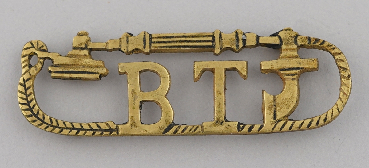 Merke eller lite skilt i gult metall. Ornamentet er forma som ein telefon (?) med bokstavane B T inni.