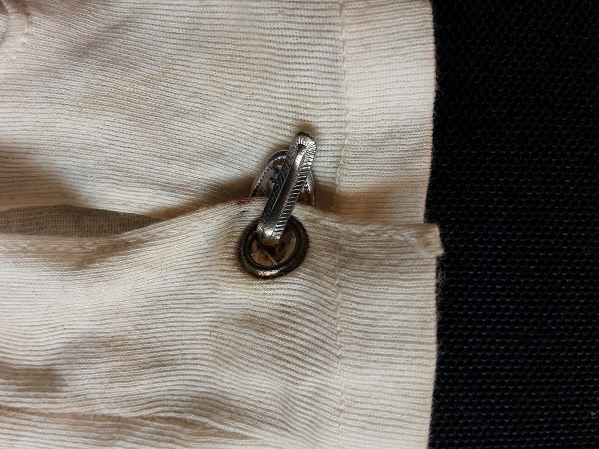 Hansker. Hvit bomull, splitt på innsiden av håndleddet som lukkes med metallspenne. Tre maskinbroderte hvite striper på handbaken.