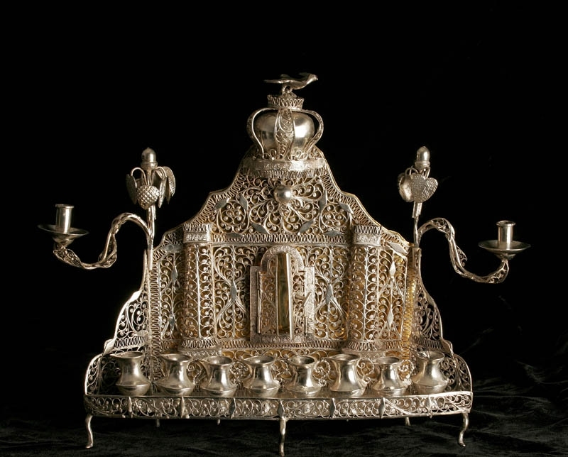 Chanukaljusstake tillverkad av silver, med åtta kannor som ljushållare. Två lösa flankerande ljushållare. Dekorerad med genombruten filigran, samt ett torahkabinett, en krona och en fågel. 

Tillverkad i Baal Shem Tov-stil.