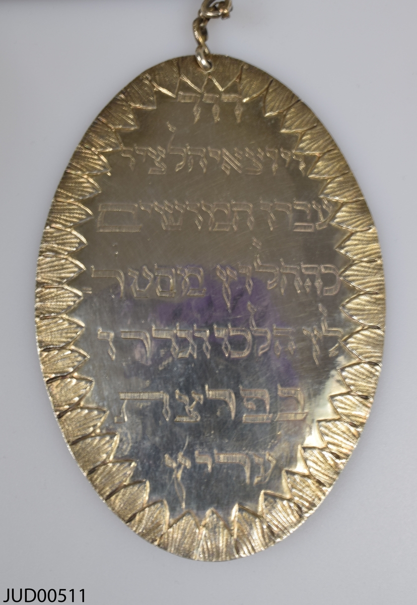 Torahsköld tillverkad av silver. Den här svenska stämplar i form av O2, kattfot, St Erik och EB. Dekorerad med lejon som flankerar en krona och sköld med hebreisk text. Tre hängande plaketter med ristad hebreisk text i nederkant.