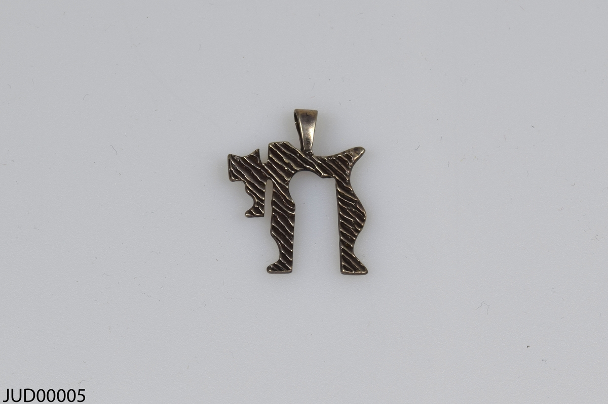 Hängsmycke tillverkat av silver i form av den hebreiska bokstaven ”Chaj” [står för liv och siffran 18]. Saknar kedja.