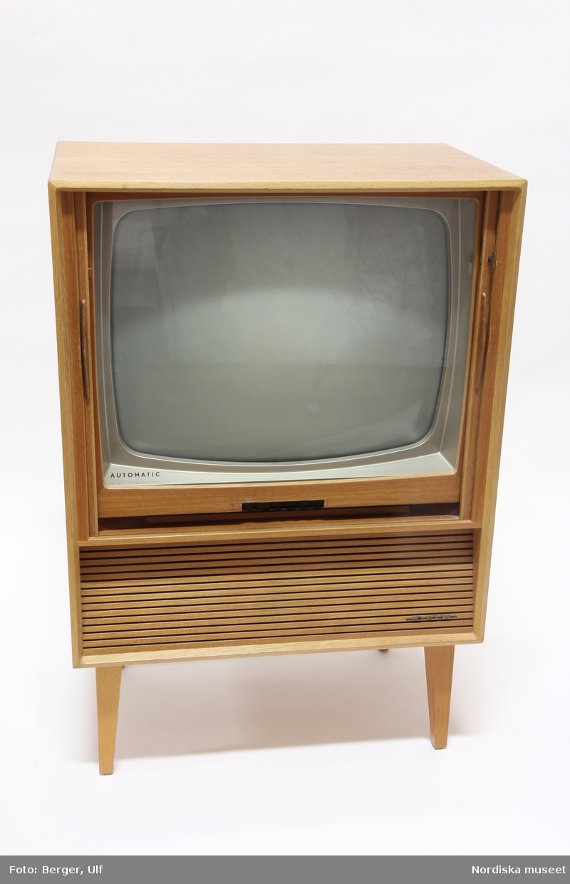 a) TV, svart/vit, av märket Dux, modell Ridå V 5910, inbyggd i teakmöbel, med 23” rör och jalusier och b:1-4) löstagbara ben. Jalusiet kan låsas med en c) nyckel av mässing. Inköpt på NK den12 december 1961.