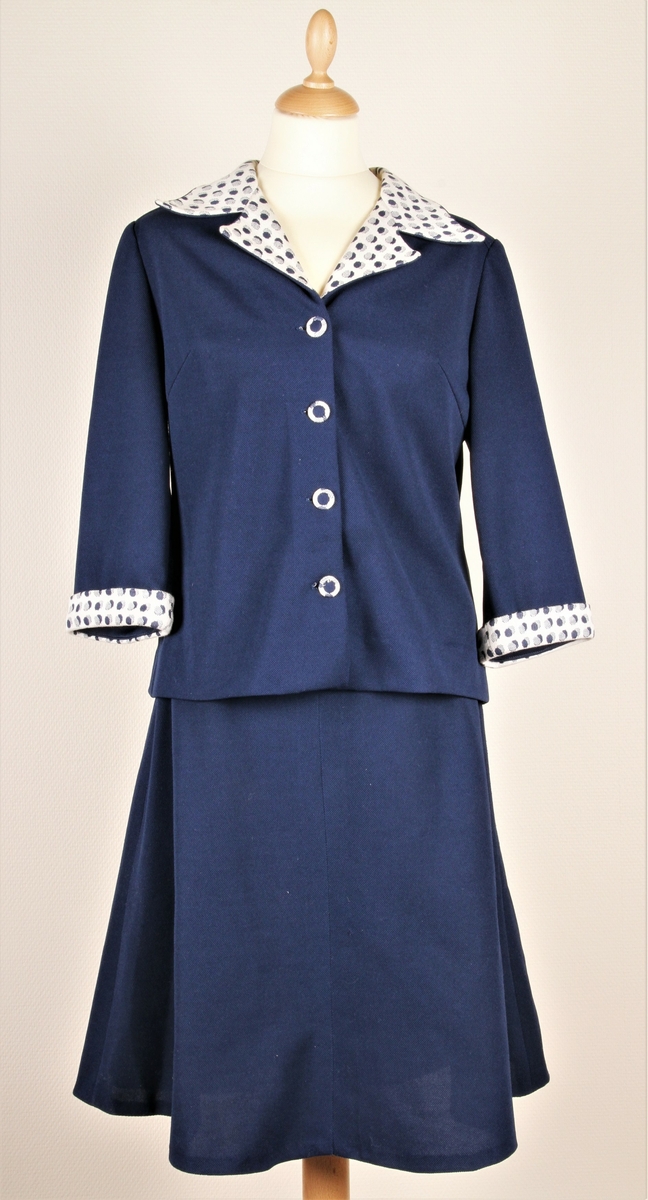 Mørk blå drakt med mønstret krage og ermkanter, jakke og skjørt. Fire knapper foran på jakken.