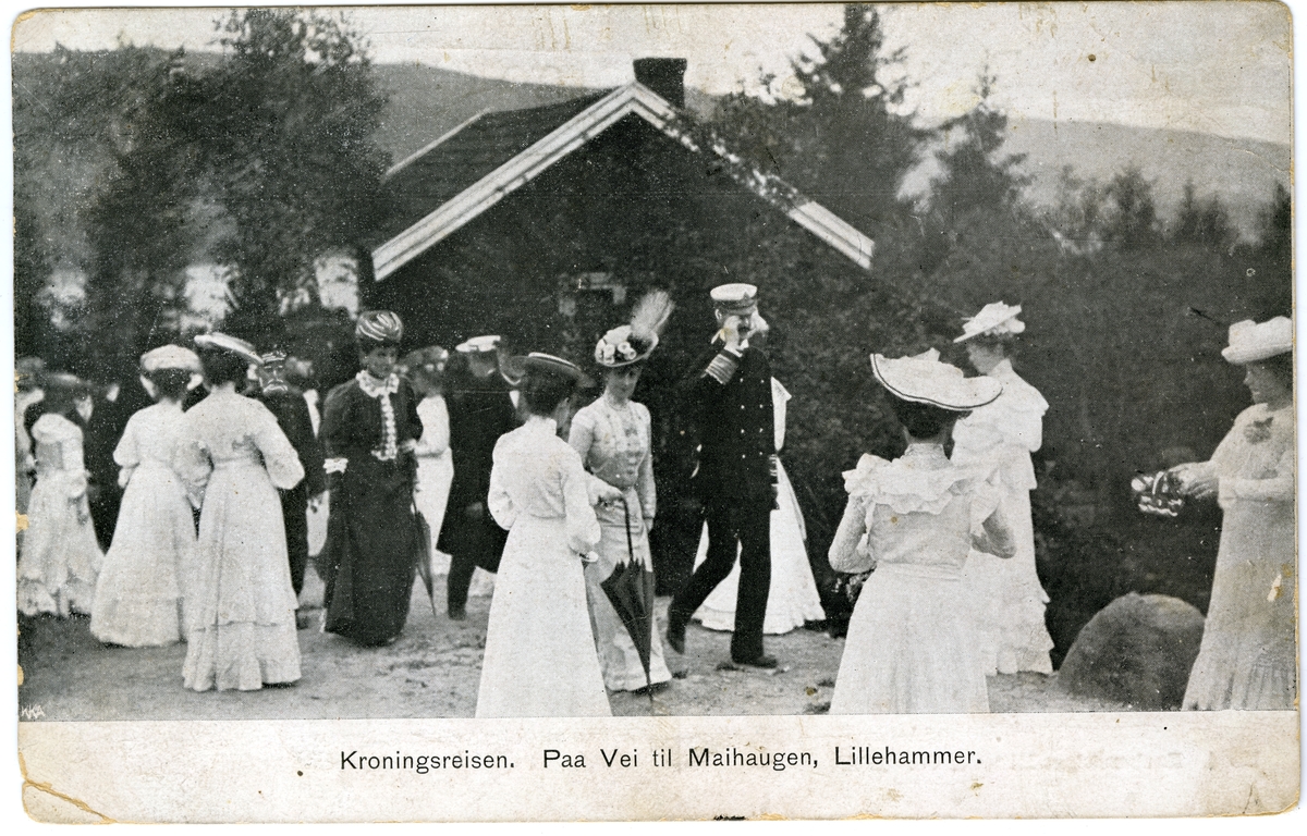 Postkort. Flere mennesker rundt kong Haakon og dronning Maud på vei til Maihaugen, Lillehammer. Kroningsreisen. Datert 02.07.1906.