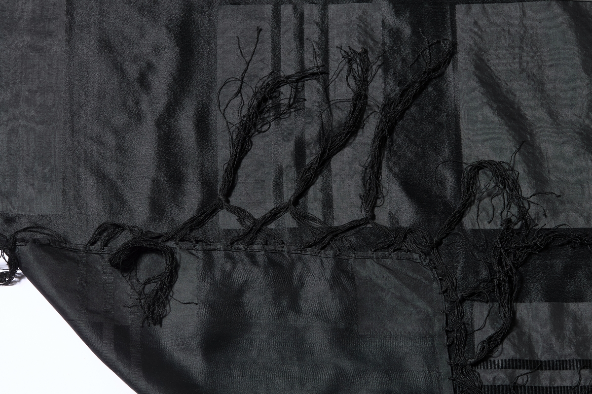 Stort, svart silkesjal med vevet mønstring av sateng i striper /ruter, samt smalere striper i ripsveving. Jarekant i to av sidene, de to andre er faldet for hånd. 