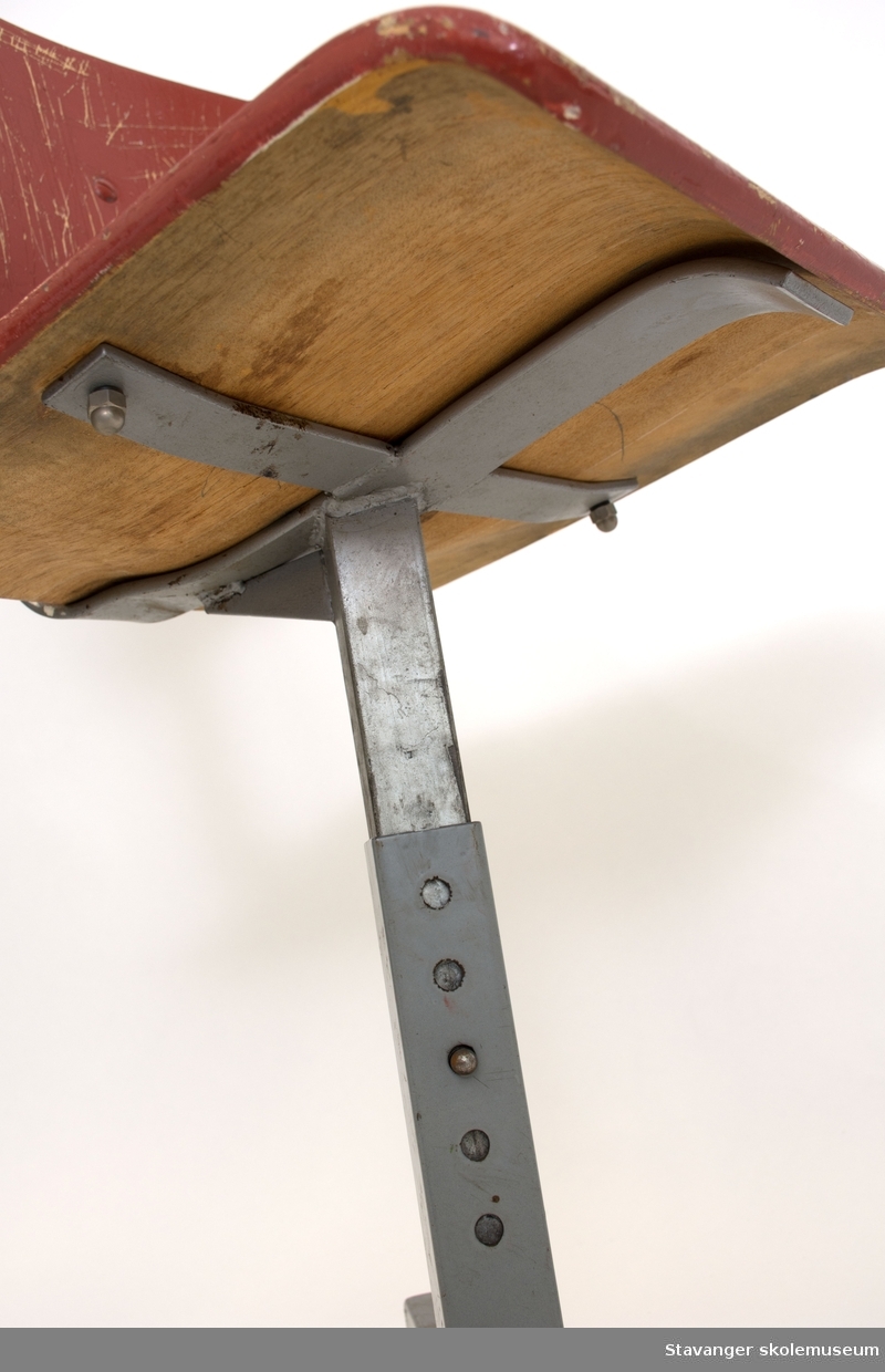 Elevstol av jern og bøyd kryssfiner. Gråmalt stål og rødmalt kryssfiner i sete og rygg. Er justerbar i høyde.