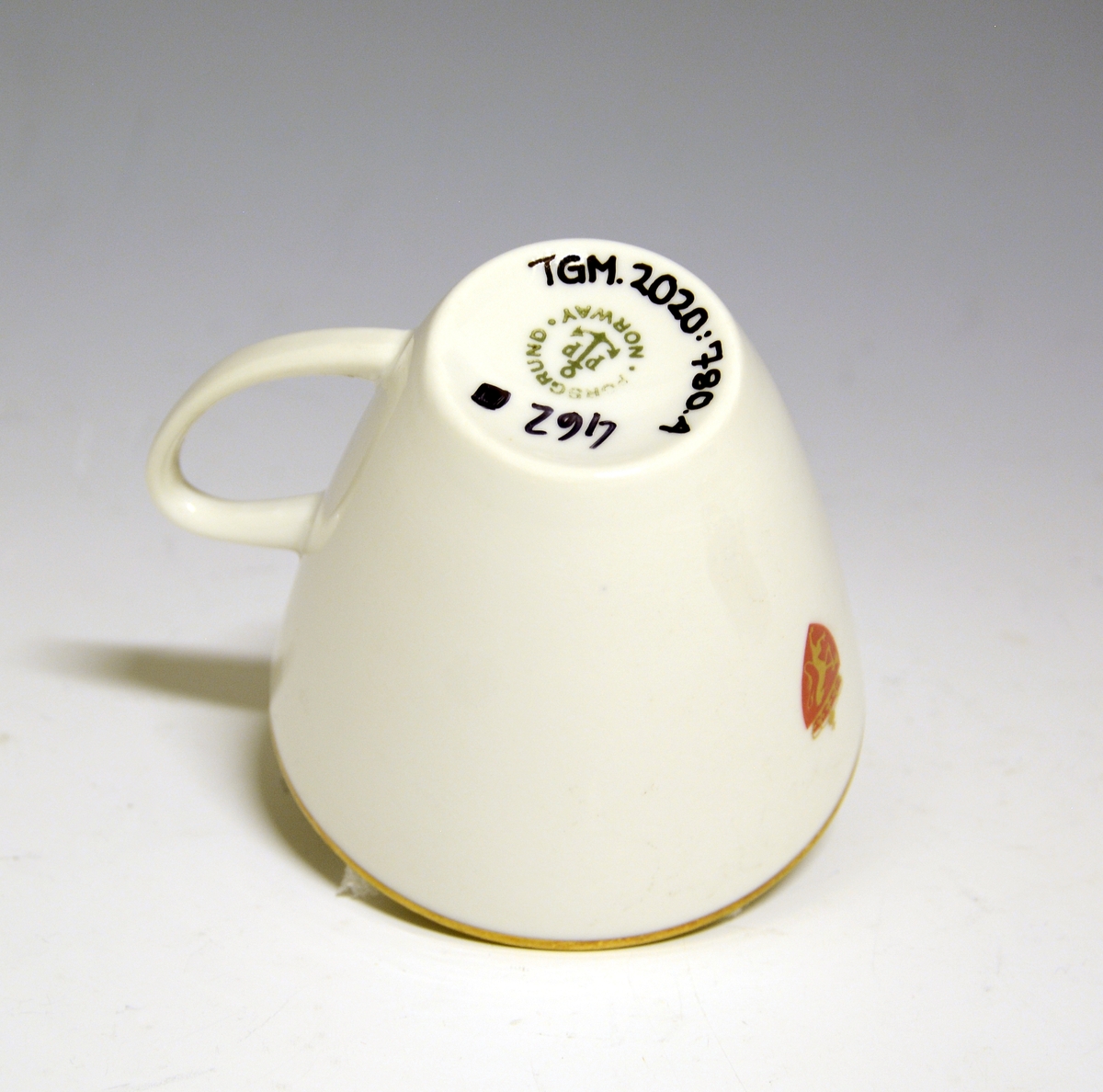 Kopp av porselen med hvit glasur. Dekorert med gullkant langs toppranden og riksvåpen.
Modell: Jubileum 2340, formgitt av Eystein Sandnes i 1959.