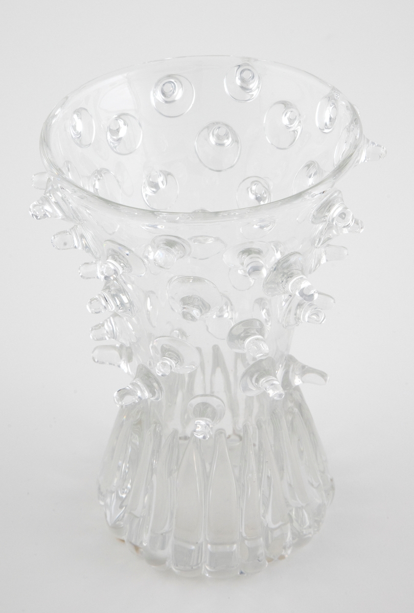 Begerformet vase i klart glass. Øvre del av er tett dekorert med plastiske "nupper" som stikker ut fra korpus, mens nedre del har dåpeformede vertikale riffler i lavt relieff.