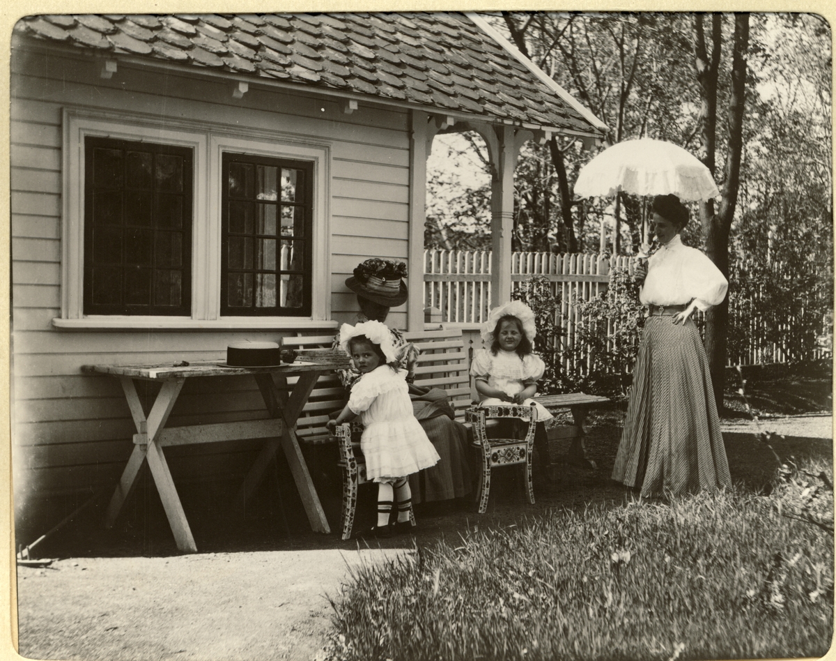 Fra hagen i Kruses gate 8 i Oslo, hjemmet til Nini og Westye Egeberg. Nini Egeberg med parasol og de to små døtrene Lucy og Mimi. På benken sitter en ukjent kvinne. Fotografert mai 1909.