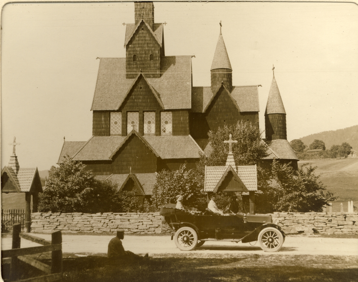 Nini Egeberg og familiens sjåfør fotografert i bilen foran Heddal stavkirke. I forgrunnen sitter en mann på plenen.