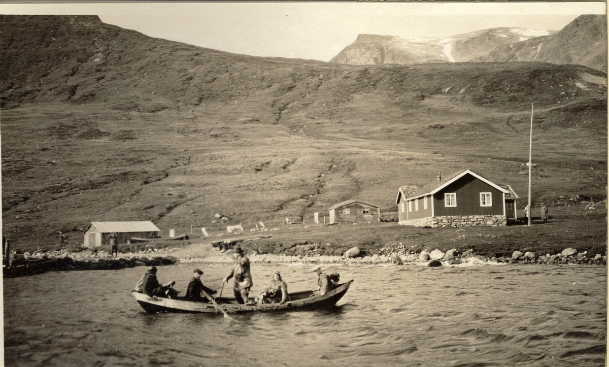 Turister og turistførere i robåt, antagelig i østenden av innsjøen Bygdin i Oppland. På land noen hytter og uthus. Fotografert 1916 under Egeberg-familiens biltur i Valdres.