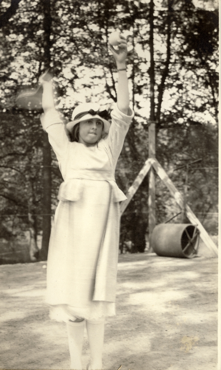 Mimi Egeberg gjør seg klar til å serve på tennisbanen. I bakgrunnen en valse til å jevne ut banen. Fotografert 1920.