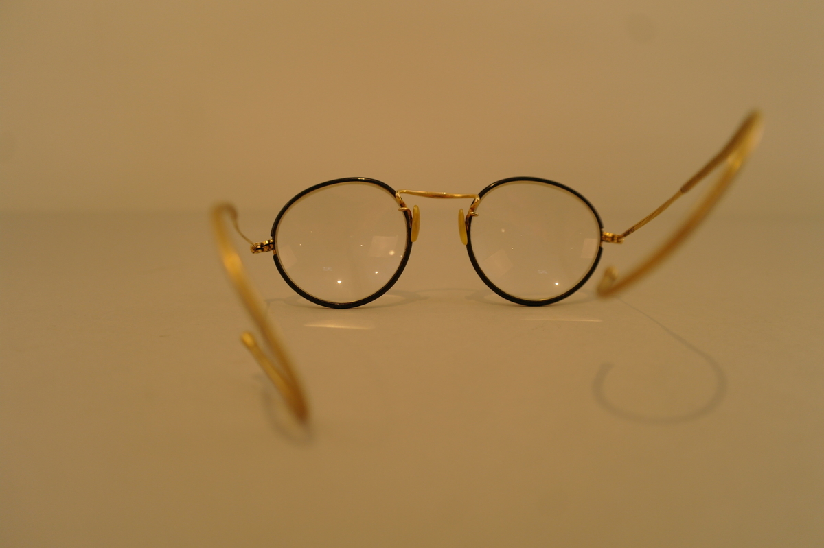 Brillehus med briller. Brilleglasene er rund og har ein svart infatning og med stenger som kan festes bak øyrane.