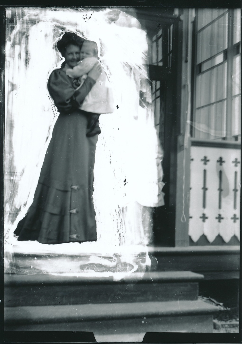 Nicoline Rydgren holder Wilhelm Fredrik, kalt Lillegutt, i armene. Kvinnen er iført lang kjole med skinkeermer. Står foran glassverandaen til bestyrervillaen på Kistefos. Hvitt felt rundt Nicoline og Wilhelm Fredrik.