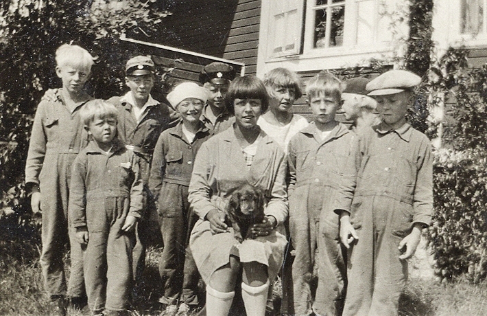 Ett gäng småpojkar och småflickor har samlats kring en större flicka med en hundvalp i knäet. 
Under fotot text: "Hällevik, 1929".