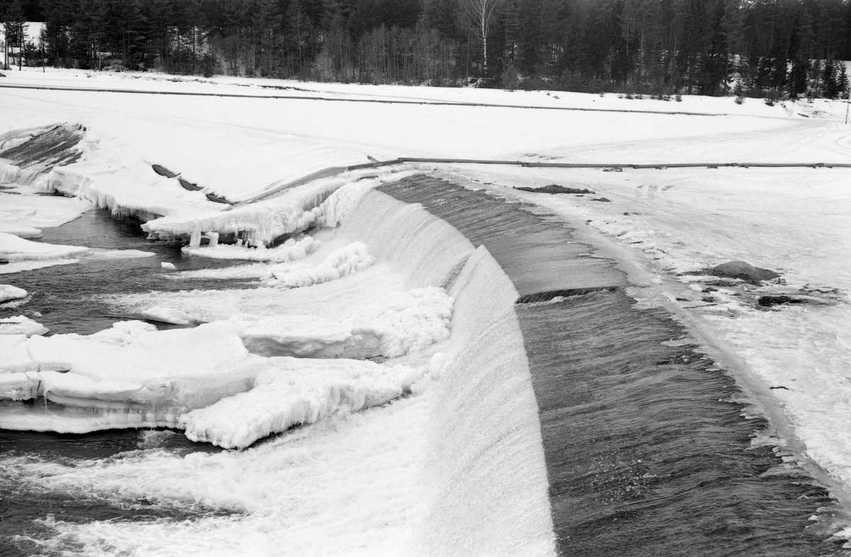 Opskuvings- eller løftedammen for kraftverksdammen ved Skjefstadfossen i Heradsbygda i Elverum, fotografert vinteren 1964.  Fotografiet er tatt fra et høytliggende punkt, antakelig en støpt plattform ved damfestet på Glommas østre bredd, rett mot den svakt buete, 312 meter lange overløpsdammen.  I forgrunnen ser vi hvordan vannet renner over damkrona i den østligste og djupeste delen av elveløpet.  Dette var for øvrig også den delen av elveløpet der fløtingstømmeret helst skulle gå.  Dette fotografiet er imidlertid tatt vinterstid, og da lå det snødekt is på den ovenforliggende kraftverksdammen og isflak i det nedenforliggende elveleiet.