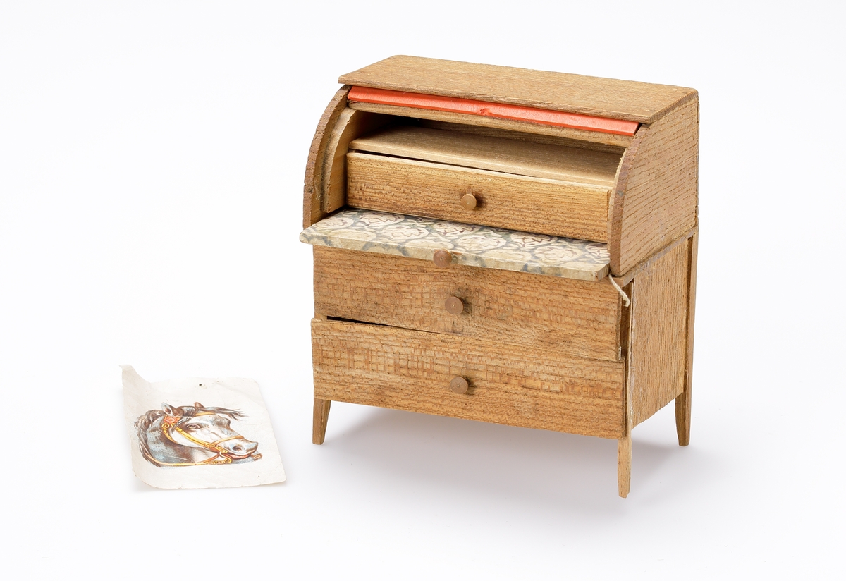Chiffonjé (dockmöbel) av brunt trä, med två lådor och uppfällbar (rull-)klaff. Bakom klaffen en låda och en hylla. Klaffen klädd med stormönstrat papper. I en låda ligger det ett bokmärke.