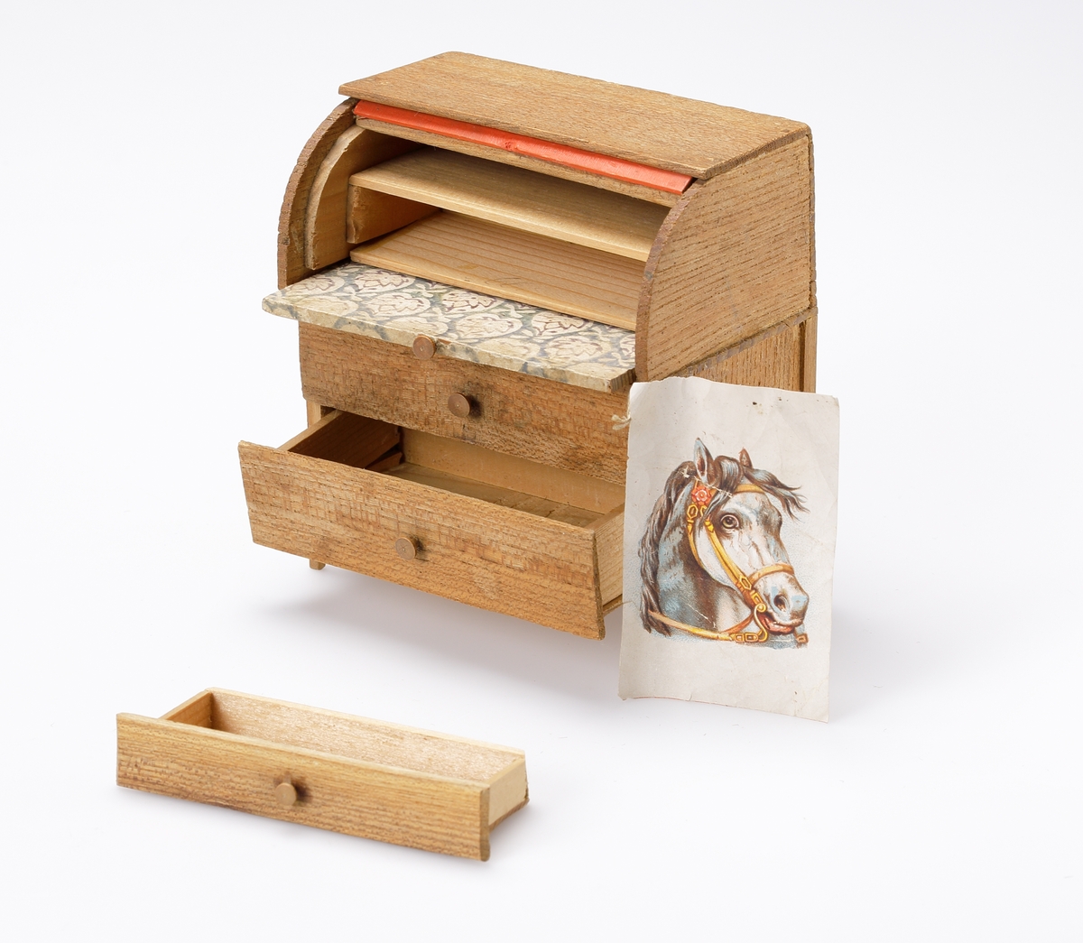 Chiffonjé (dockmöbel) av brunt trä, med två lådor och uppfällbar (rull-)klaff. Bakom klaffen en låda och en hylla. Klaffen klädd med stormönstrat papper. I en låda ligger det ett bokmärke.