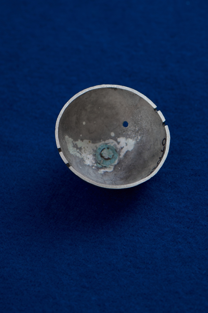 En silverplåt formad rund och skålformad. Silverplåten har en knopp upptill och hål på sidan. Fyra fördjupningar är gjorda längs kanten. Plåten är kraftigt oxiderad.
