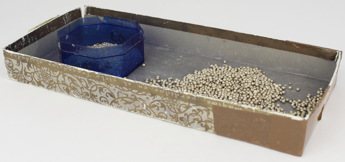 Ett föredetta lock som används till en låda. Lådan är tejpad med brun tejp och sliten i hörnen. Ytan är lilaaktig med slinger- och blommönster i silverfärg.