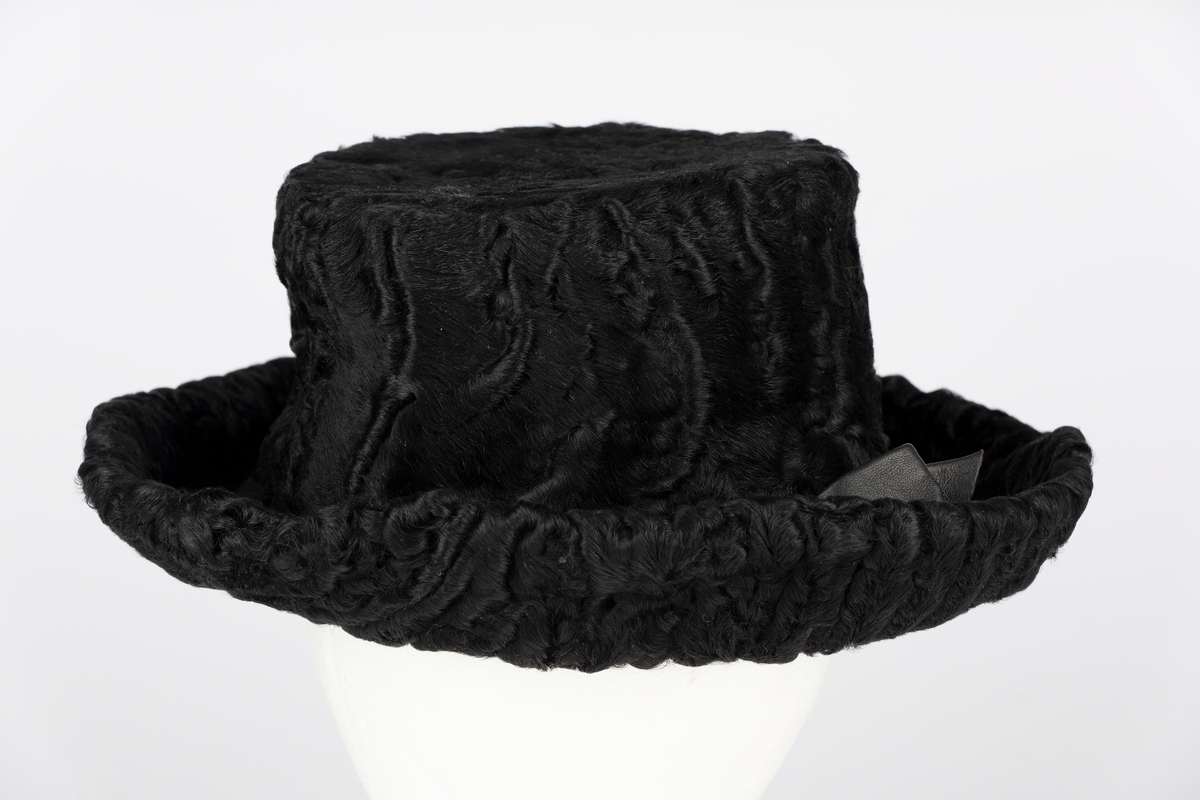 Hattepull i persianerskinn. Bremi sort filt trukket på undersiden med sort persianerskinn. Pyntesløyfe i skinn.