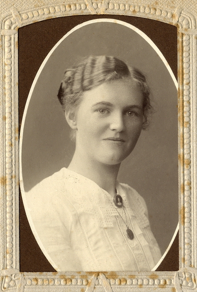 En ung kvinna i ljus blus med en brosch i halsgropen och halskedja med medaljong. 
Bröstbild, halvprofil.