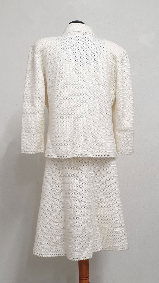 Hvit heklet drakt bestående av ermeløs kjole og jakke. Jakken har knapper i front, pyntelommer på hvert forstykke, skjortekrage og skulderputer. Flekk i front på både jakke og kjole.