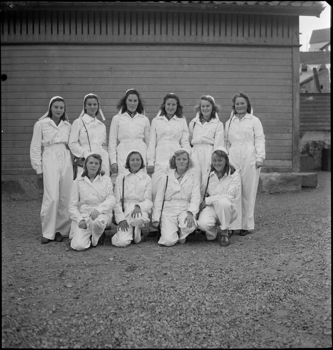 En gruppe med unge kvinner i hvit uniform. På armen har de symbolet til kong Håkon samt et kors med anker i et hjerte. De har hvite luer og vesker.